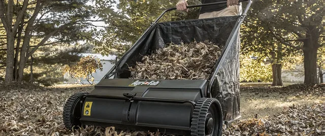 A person using a push lawn sweeper in a yard filled with fallen leaves