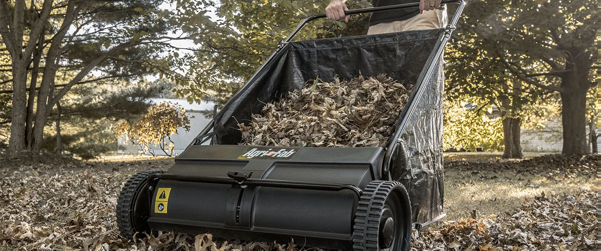 A person using a push lawn sweeper in a yard filled with fallen leaves