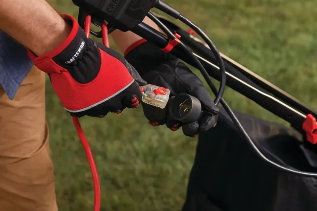Close-up of a person wearing red and black gloves plugging a power cord into a CRAFTSMAN electric lawn mower. The mower is outdoors on green grass.