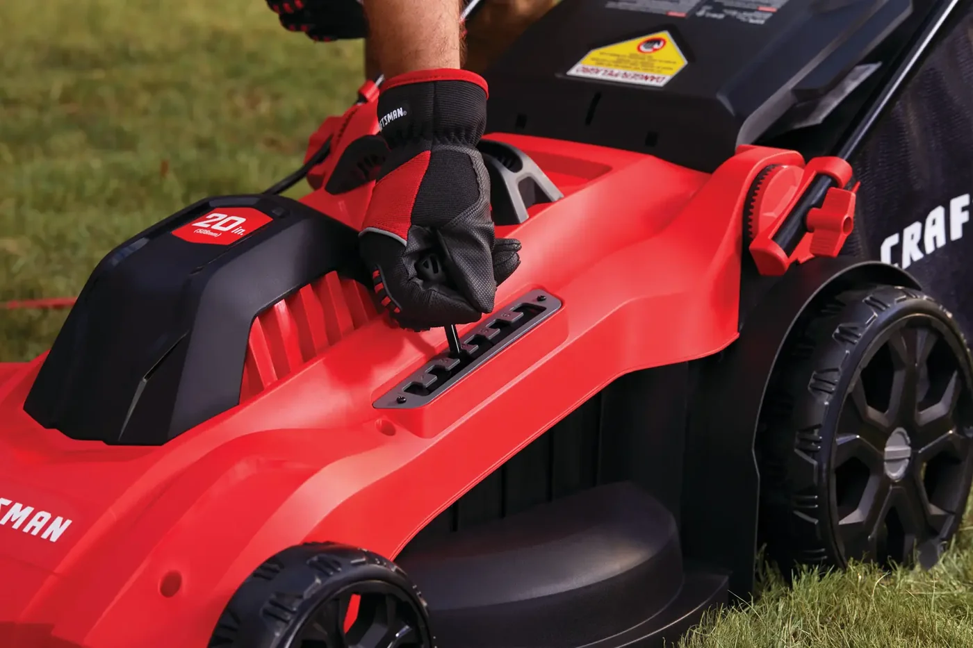 Close-up of a person wearing red and black gloves adjusting the height setting lever on a red CRAFTSMAN corded electric lawn mower, shown on grass.
